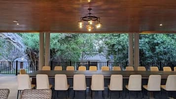 Un espace de restauration intérieur avec une longue table et des chaises, un plafond en bois et une vue sur les arbres à l’extérieur.