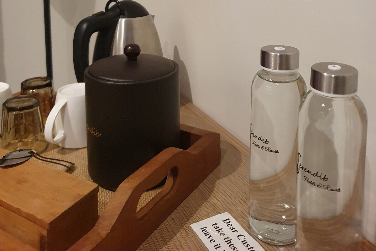 A wooden tray with a black pot, two glass bottles, a white mug, and a black kettle on a table.