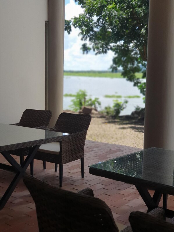 A patio with wicker furniture and a glass table, overlooking a body of water and greenery. - Waters Edge By Serendib (Liwonde)