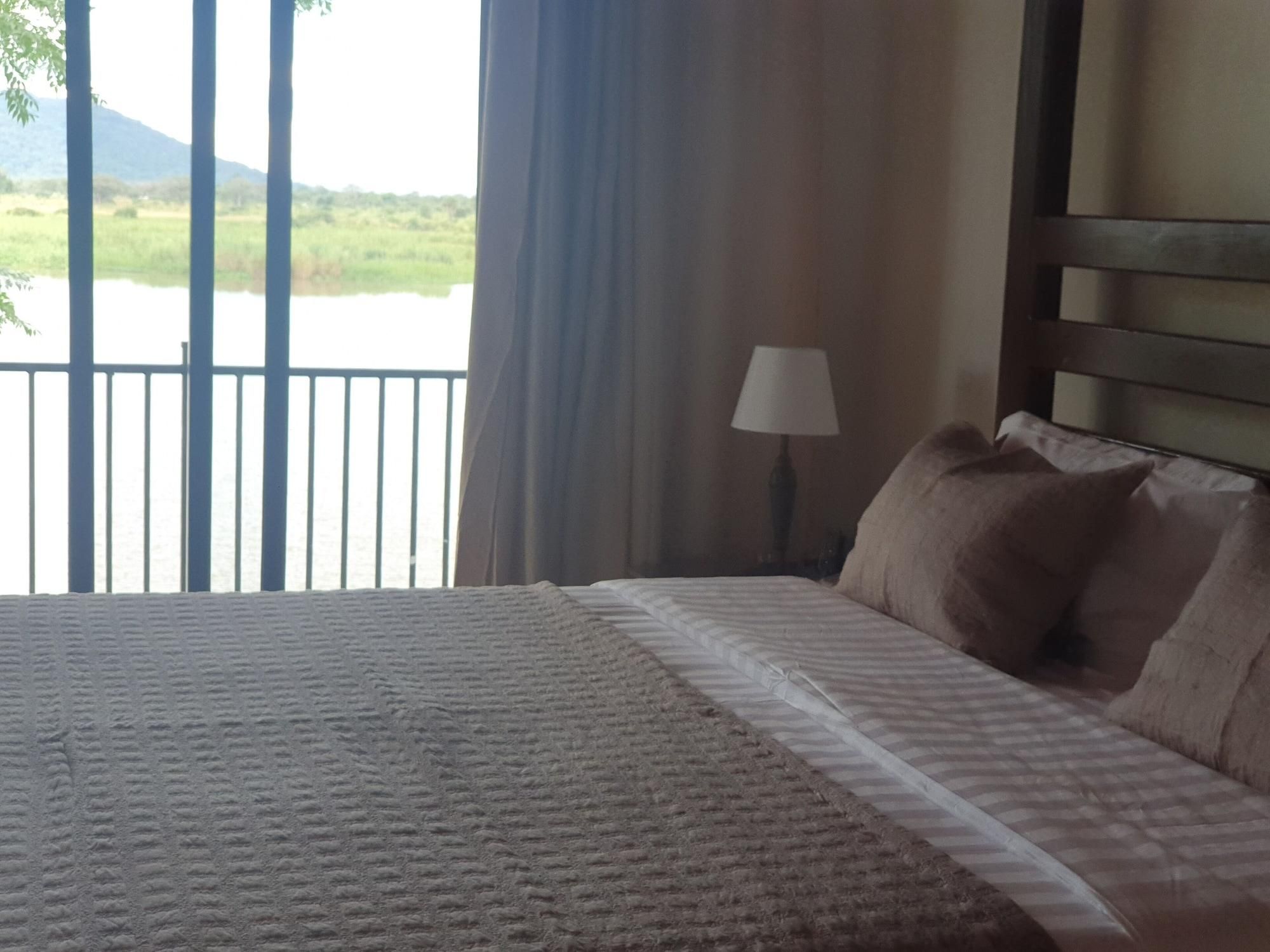 A bed with a patterned bedspread, a lamp, and a view of a body of water and greenery through a window.