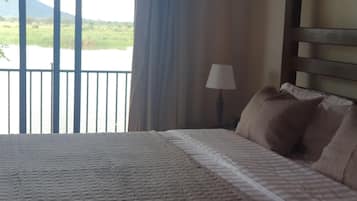 A bed with a patterned bedspread, a lamp, and a view of a body of water and greenery through a window.