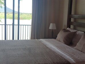 Una cama con una colcha estampada, una lámpara y vistas a un cuerpo de agua y vegetación a través de una ventana.