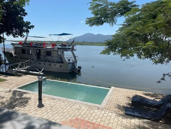A boat docked by a calm lake with a small pool and lounge chairs. - Waters Edge By Serendib (Liwonde)