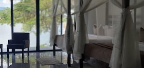 A bedroom with a bed, a chair, and a view of the sea through large windows. - Waters Edge By Serendib (Liwonde)