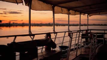 A boat deck with a covered seating area, a metal railing, and a view of a sunset over a body of water.