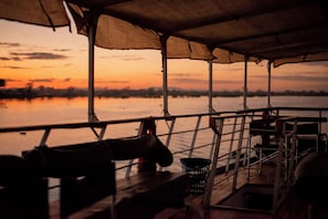 Cubierta de barco con zona de estar techada, baranda de metal y vista del atardecer sobre el agua.