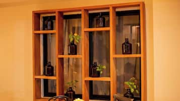 A dining area with a wooden table and chairs, a window with shelves holding bottles and plants, and a power outlet on the wall.