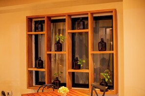Un comedor con mesa y sillas de madera, una ventana con estantes que sostienen botellas y plantas, y un enchufe en la pared.