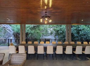 A covered outdoor dining area with a long table and chairs, a chandelier, and a view of trees and a building in the background. - Waters Edge By Serendib (Liwonde)