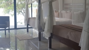 A bedroom with a bed under a canopy, a chair, and a view of trees through large windows. - Waters Edge By Serendib (Liwonde)