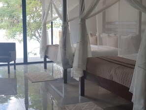 A bedroom with a bed under a canopy, a chair, and a view of trees through large windows. - Waters Edge By Serendib (Liwonde)