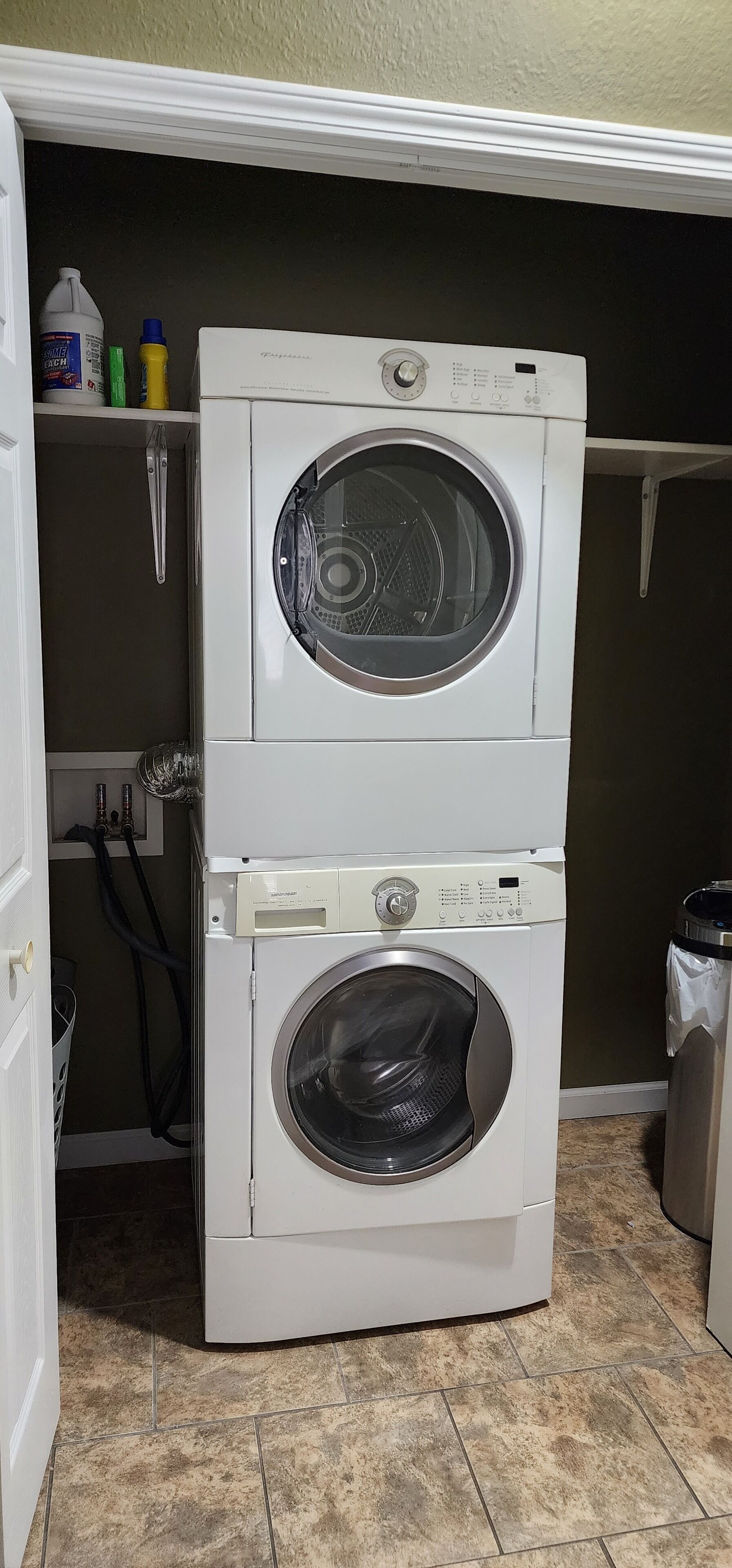 A stacked washer and dryer unit in a laundry room with a tiled floor and a shelf above holding cleaning supplies.