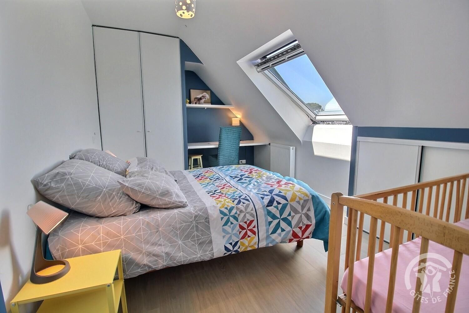 Ein Schlafzimmer mit schrägem Dach, einem Oberlicht, einem Bett mit gemusterten Textilien, einem Nachttisch und einem Babybett.