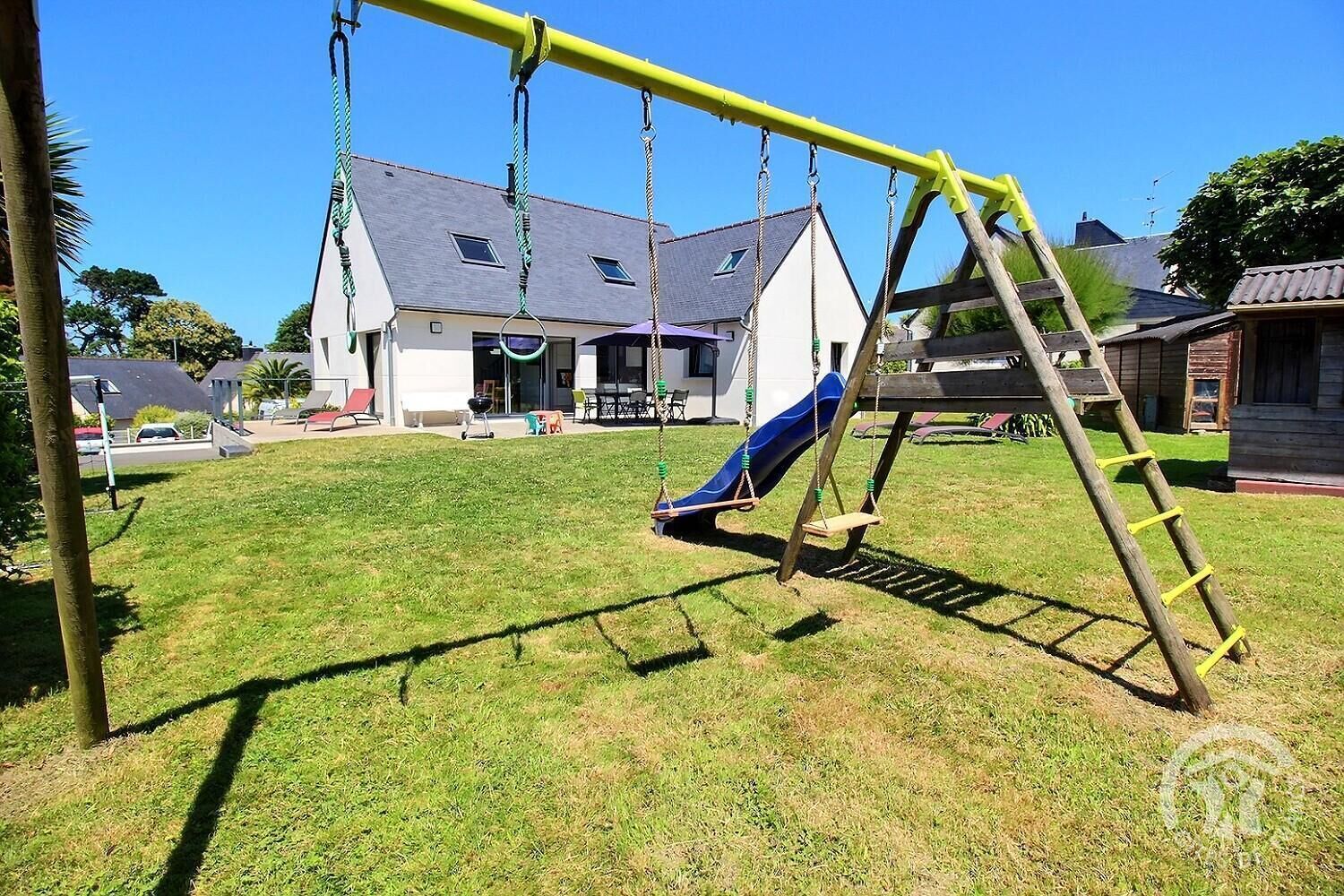 Ein Spielplatz mit Schaukeln und Rutsche, umgeben von Rasen und Häusern im Hintergrund.