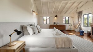 Un dormitorio con una cama grande, mesita de noche, lámpara y vistas al exterior a través de ventanas.