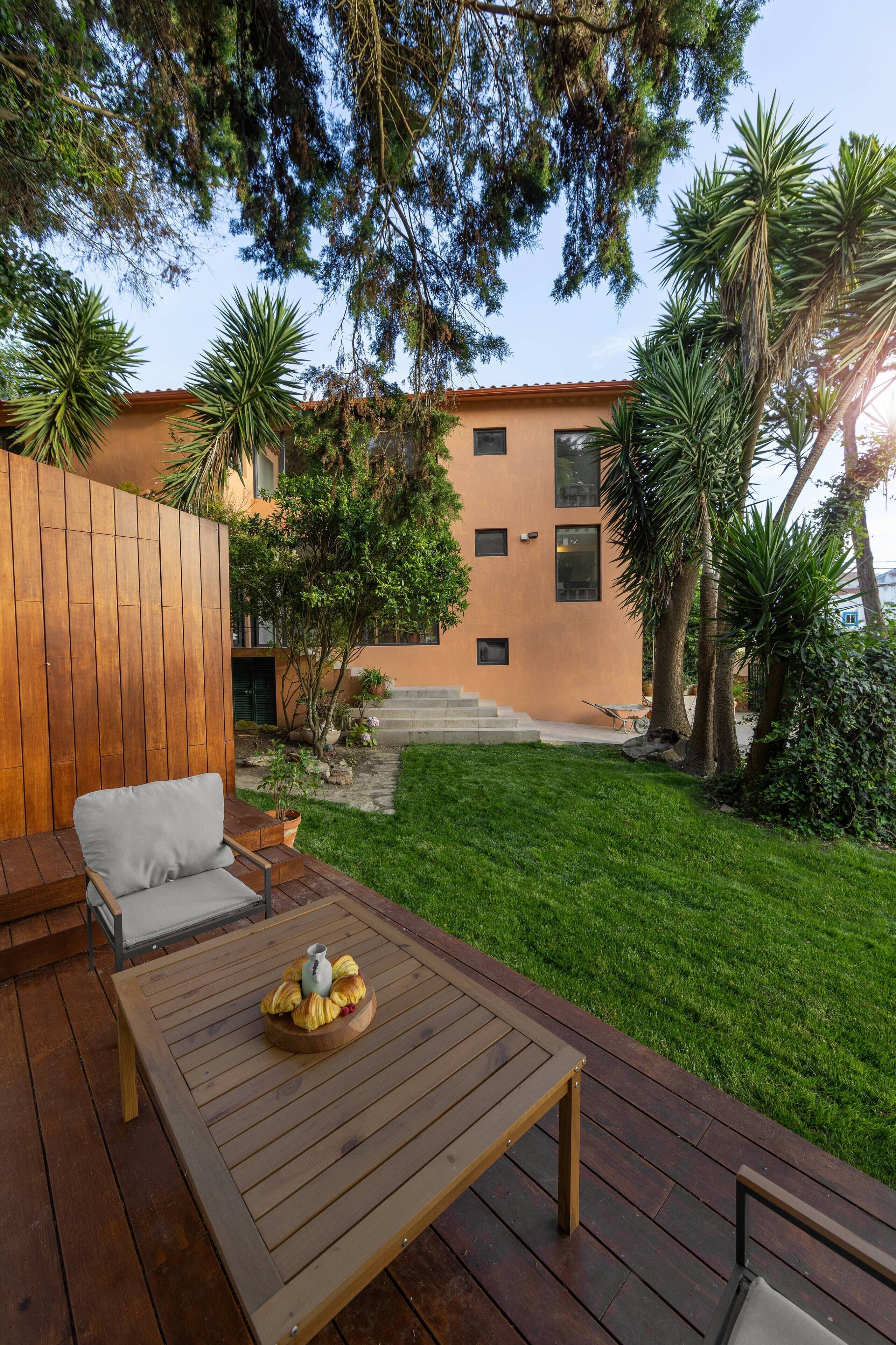 A wooden deck with a table and chairs, a small garden with grass and trees, and a building with a red roof in the background.