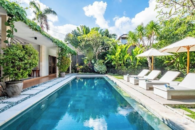 A swimming pool with lounge chairs and umbrellas, surrounded by lush greenery and a clear sky.