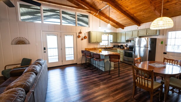 A spacious kitchen with wooden floors, a dining area with a wooden table and chairs, and a built-in island with bar stools. - “The Sugarloaf Mountain Cabin” A Charming Lakefront Escape!! (Higden)