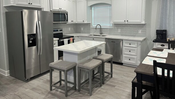 A modern kitchen with white cabinetry, stainless steel appliances, a central island with stools, and a dining area with a dark wooden table and chairs. - Brand new beach home 3BR near beach and fishing (Surfside Beach)