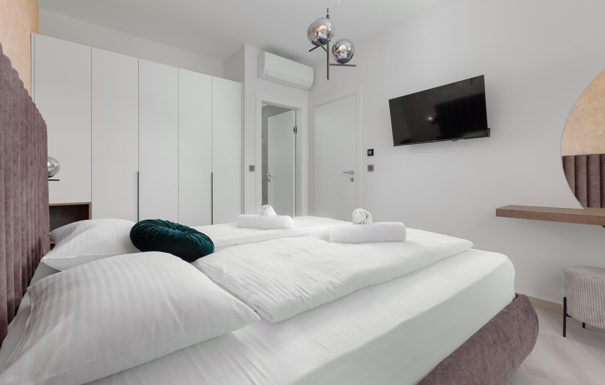 A modern bedroom with a large bed, white walls, a flat-screen TV, and a built-in air conditioning unit.