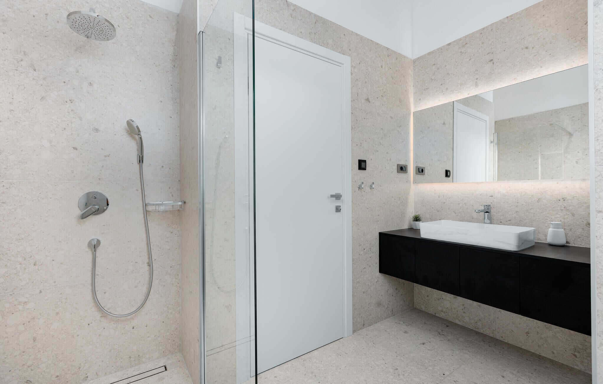Un baño moderno con cabina de ducha de vidrio, una puerta blanca, un mueble de tocador negro con lavamanos blanco y un espejo grande sobre el mueble.