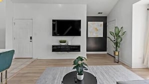 Una sala de estar moderna con un televisor de pantalla plana montado en la pared, un mueble de entretenimiento negro debajo, una mesa de centro redonda con una planta, y una planta grande en maceta al lado.