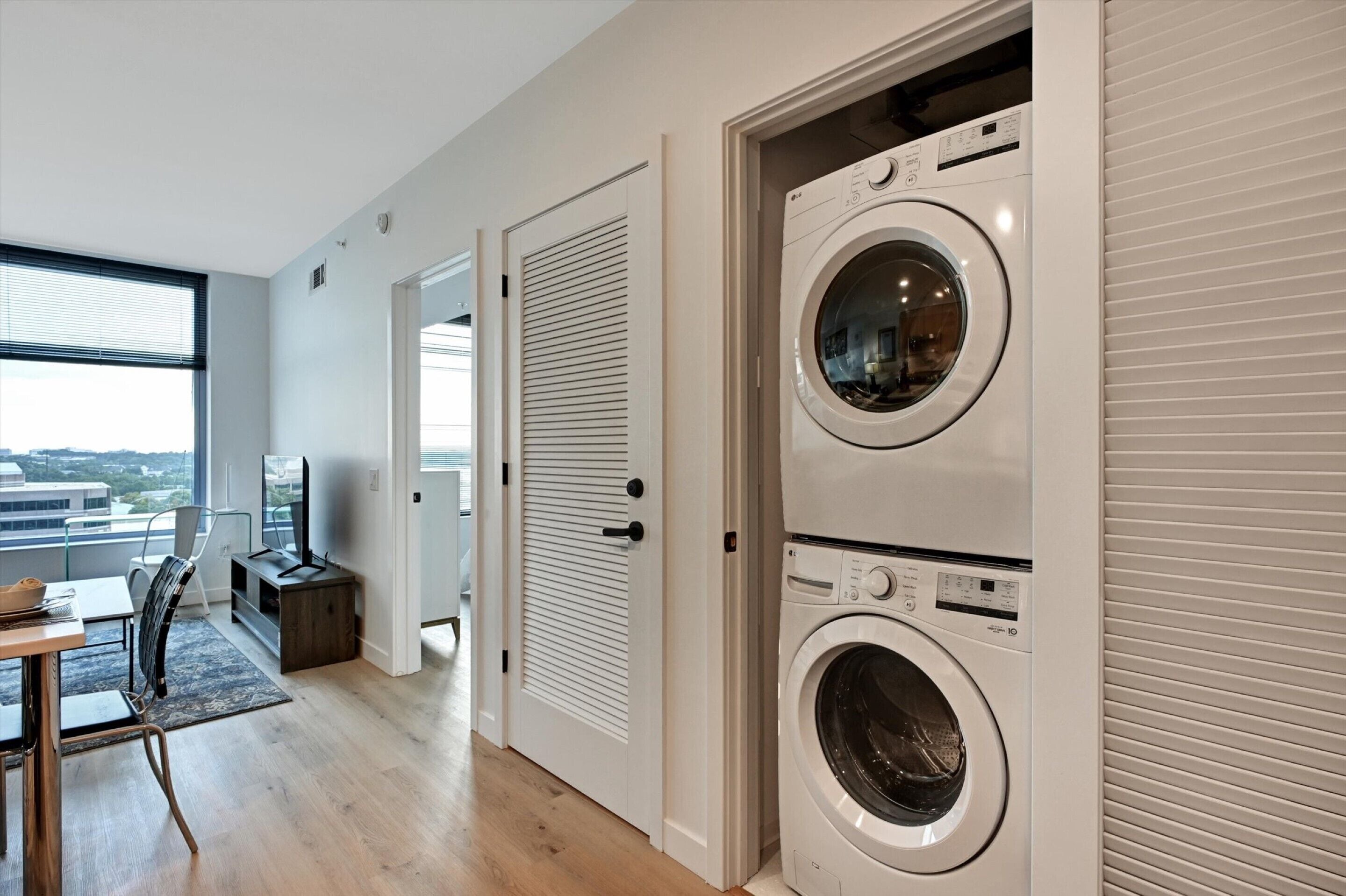 A modern laundry room with two stacked washing machines, a dining area with a table and chairs, and a large window offering a city view.