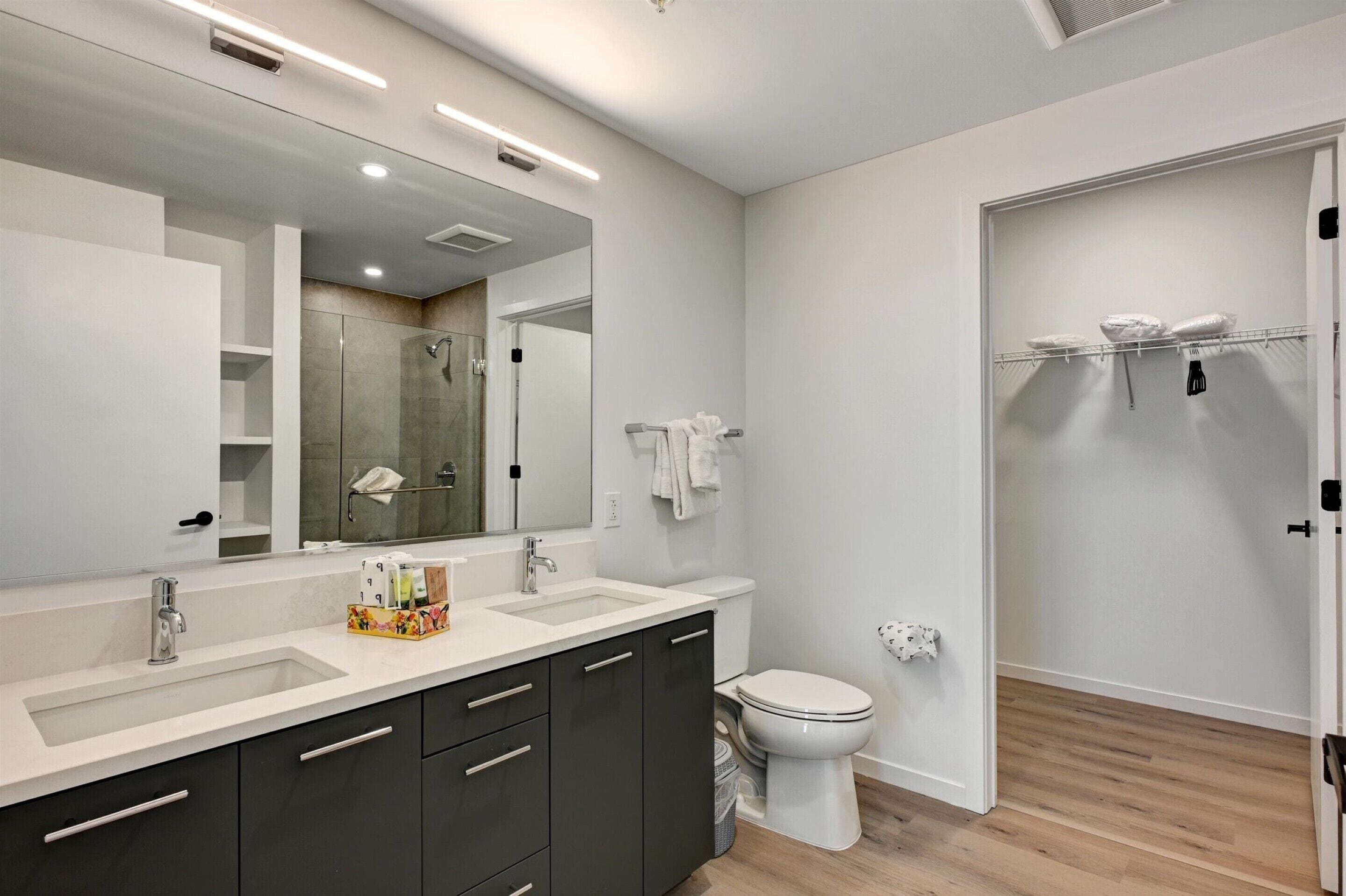 A modern bathroom with a large mirror, two sinks, a toilet, and a shower area.