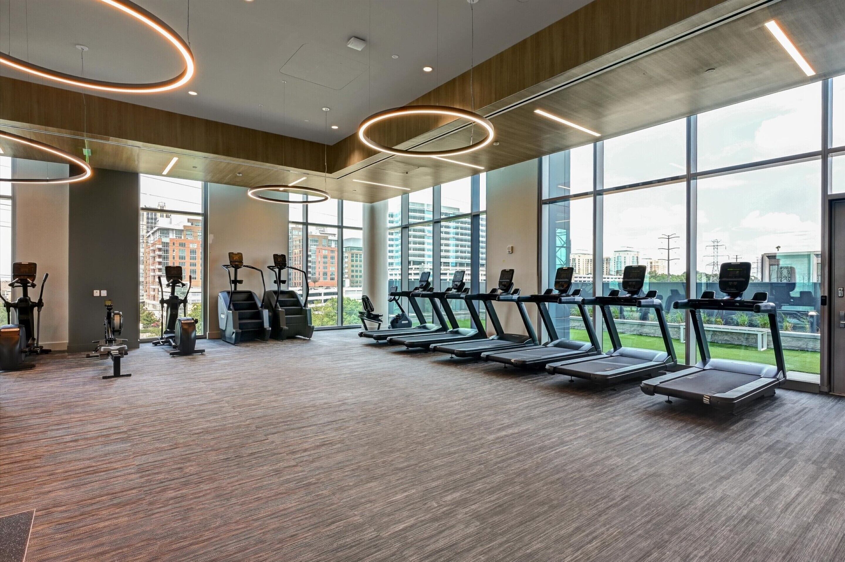 A modern gym with treadmills, ellipticals, and a large window offering a city view.