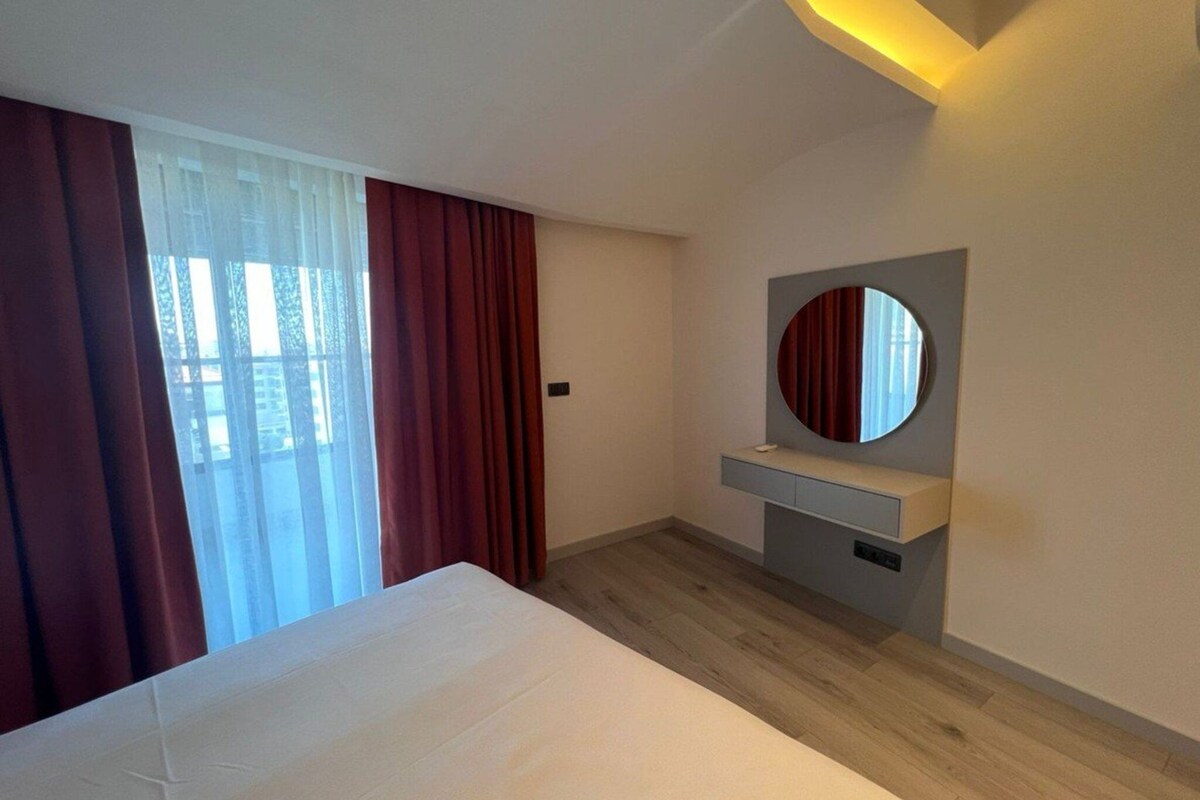 A modern hotel room with a large bed, a bedside table, a mirror, and a view of the outdoors through a window with curtains.