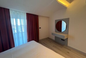 A modern hotel room with a large bed, a bedside table, a mirror, and a view of the outdoors through a window with curtains. - Ulu Residence Alanya (Alanya)