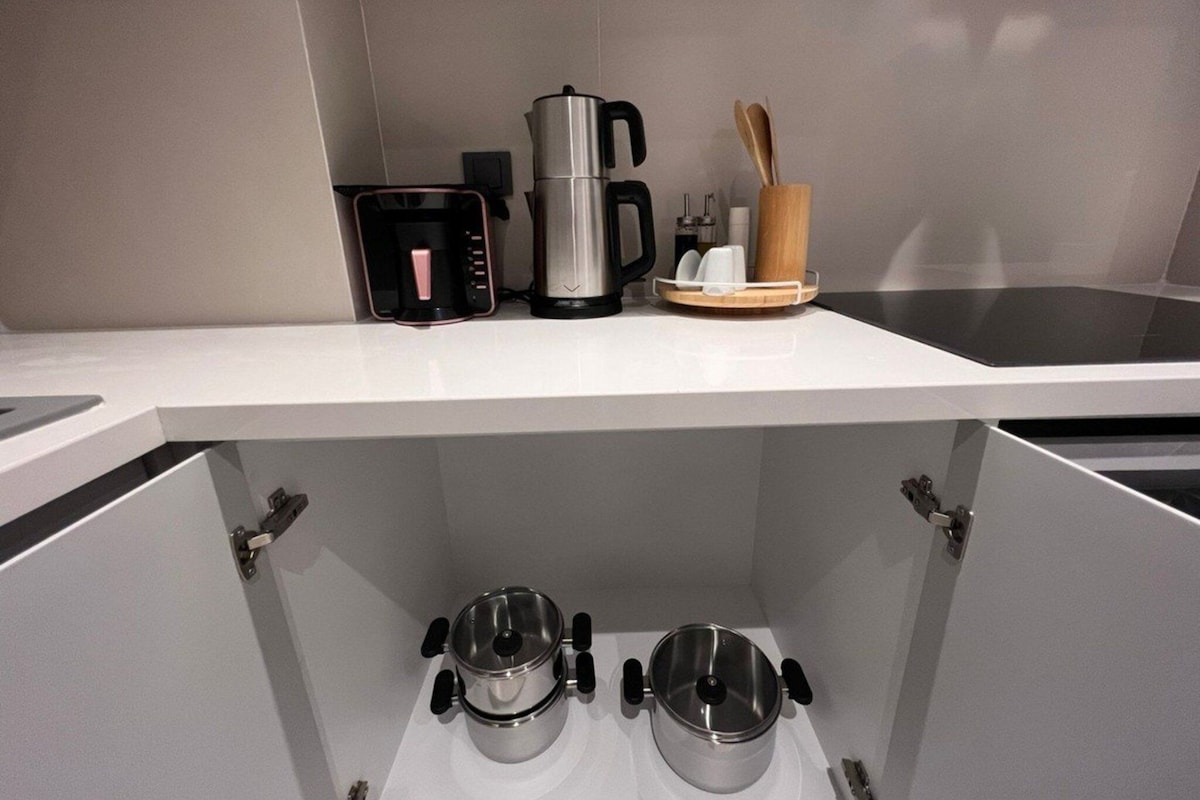 A kitchen with a white countertop, a coffee maker, a kettle, and a wooden utensil holder. There are two stainless steel pots on the lower shelf.