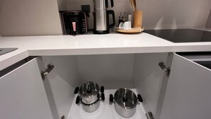 Una cocina con encimera blanca, cafetera, hervidor y un utensilio de madera. Hay dos ollas de acero inoxidable en el estante inferior.
