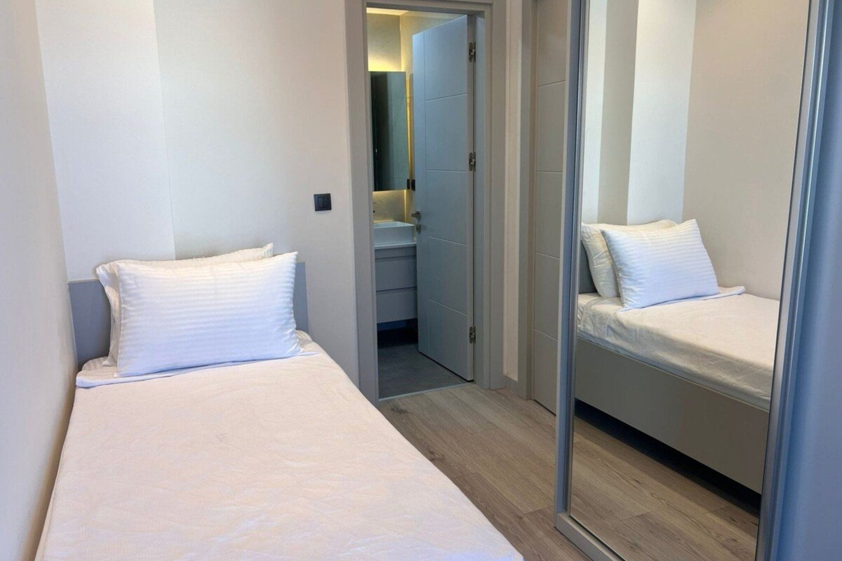 Ein schmales Hotelzimmer mit zwei Einzelbetten, einem Spiegel und einem kleinen Badezimmer, das durch eine offene Tür sichtbar ist.