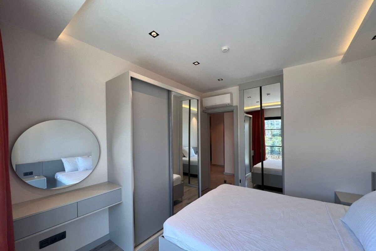 A modern bedroom with a large bed, a bedside table, a mirror, and a wardrobe.