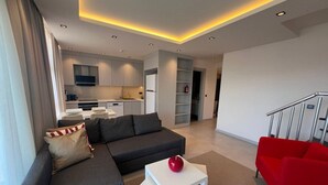 Ein modernes Wohnzimmer mit einem grauen Sofa, einem roten Sessel und einem weißen Couchtisch. Im Hintergrund ist eine Küche mit weißen Schränken und einem eingebauten Backofen zu sehen.