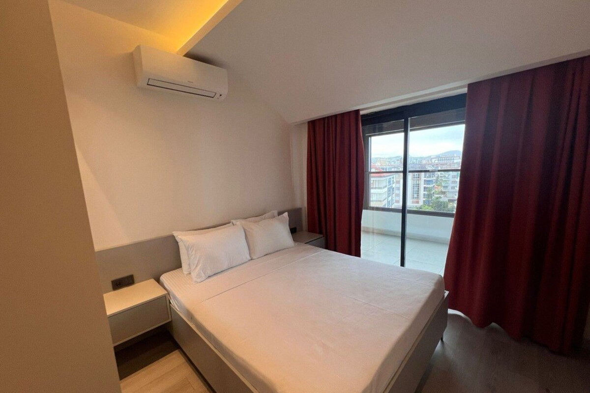 A hotel room with a large bed, a nightstand, a built-in air conditioning unit, and a view of the city through a large window with red curtains.