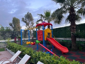 A playground with a red slide and climbing structures, surrounded by palm trees and a hedge. - Ulu Residence Alanya (Alanya)