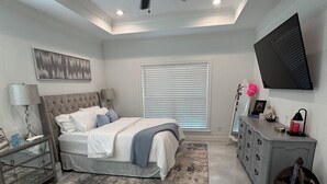 Een slaapkamer met een bed, een plafondventilator, een televisie, een raam met jaloezieën en een dressoir.