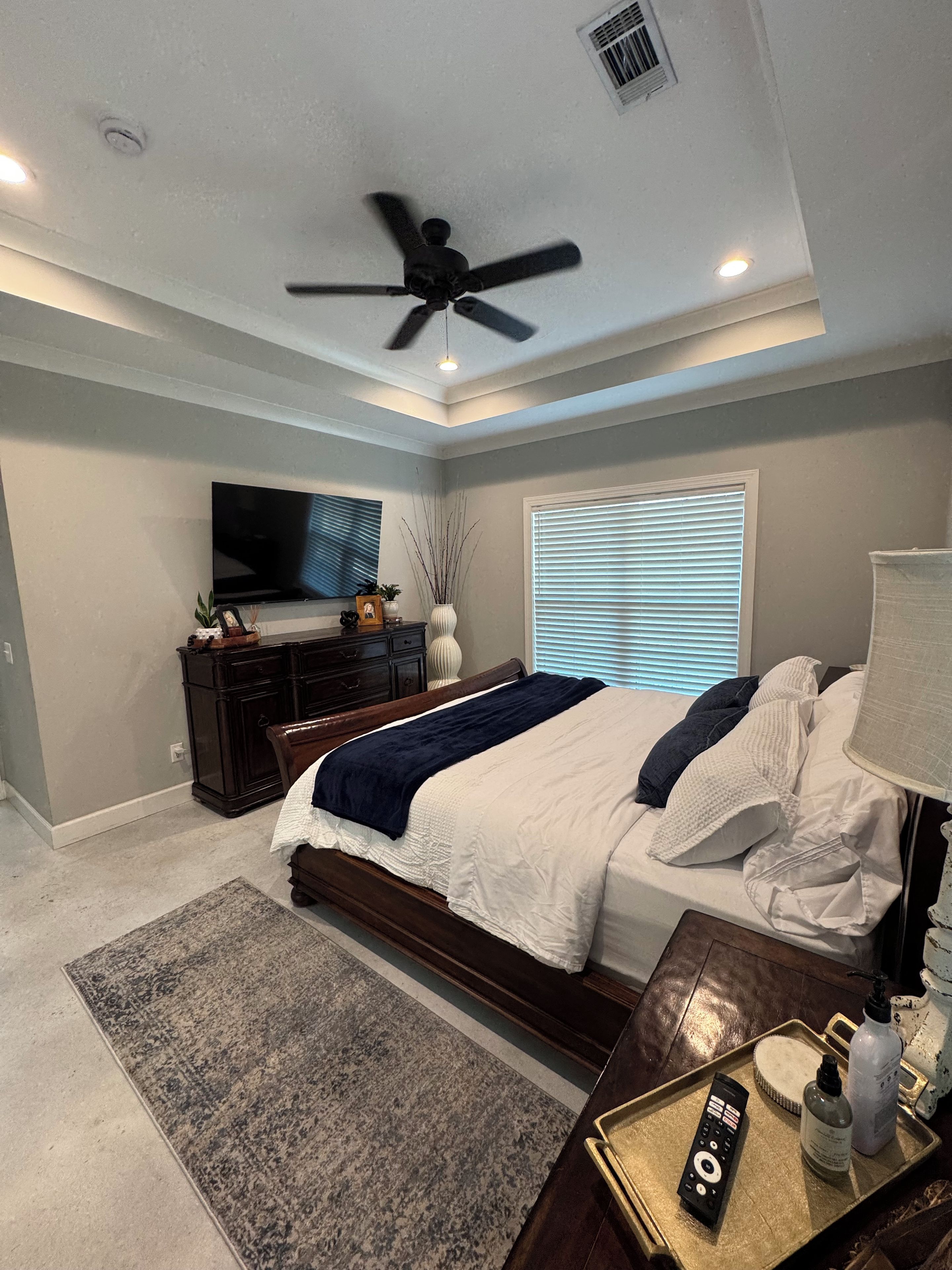 A bedroom with a bed, a TV, a ceiling fan, and a window with blinds.