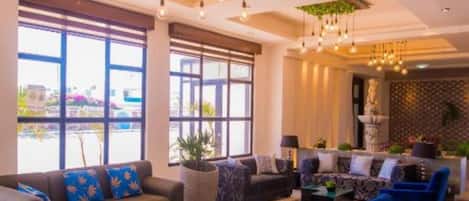 A spacious living area with a sofa set, armchairs, and a coffee table. Large windows provide natural light and views of the outside.