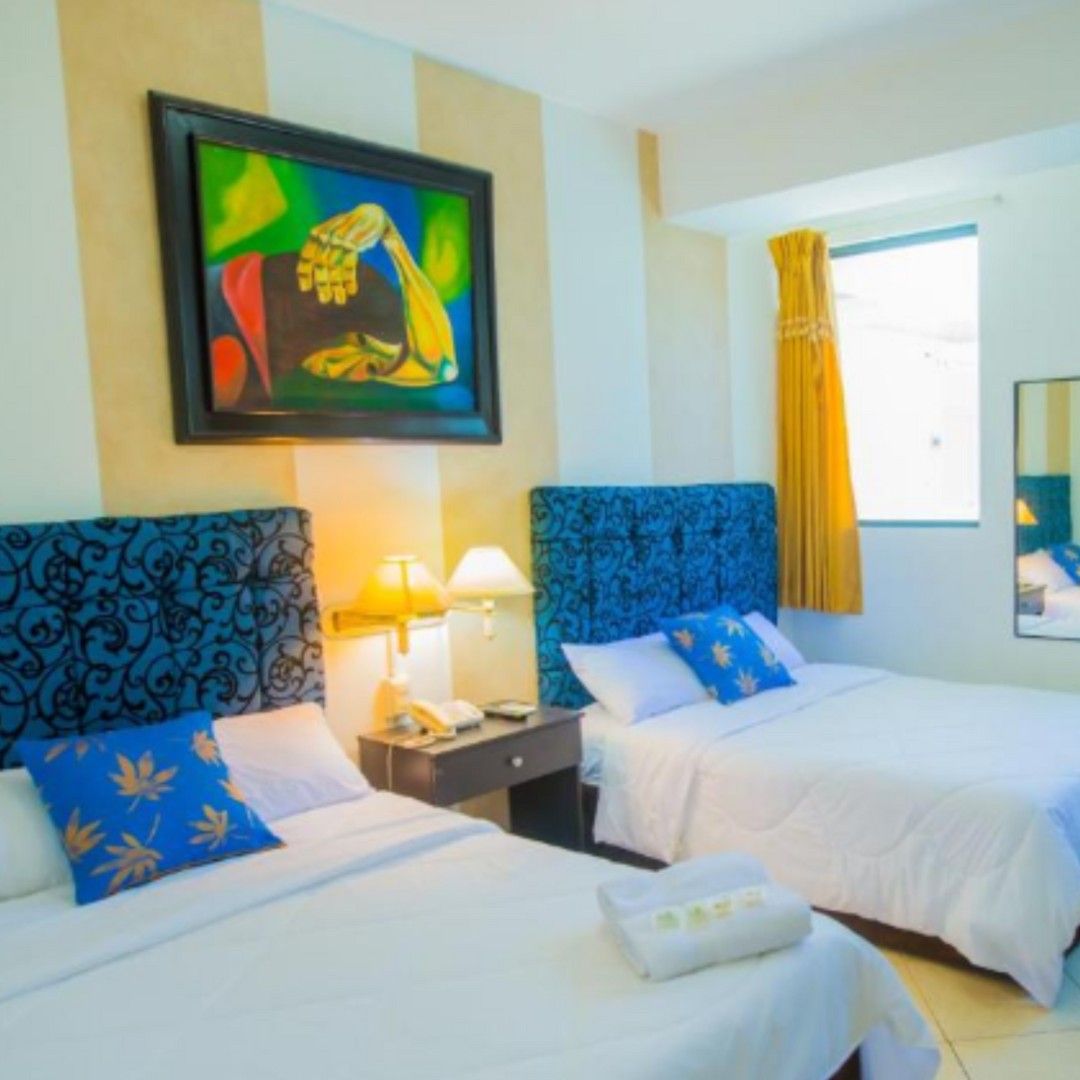 Ein Hotelzimmer mit zwei Betten, beide mit blauen Kopfteilen und weißer Bettwäsche. Über den Betten hängt ein Gemälde, ein Fenster mit gelben Vorhängen und ein Spiegel an der Wand sind ebenfalls vorhanden.