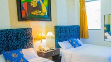 Ein Hotelzimmer mit zwei Betten, beide mit blauen Kopfteilen und weißer Bettwäsche. Über den Betten hängt ein Gemälde, ein Fenster mit gelben Vorhängen und ein Spiegel an der Wand sind ebenfalls vorhanden.