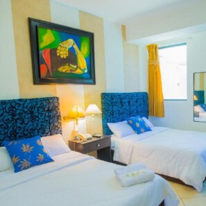 A hotel room with two beds, each with a blue headboard and white bedding. There is a painting on the wall above the beds, a window with yellow curtains, and a mirror on the wall. - Hotel Presidente Premium Salinas (Salinas)