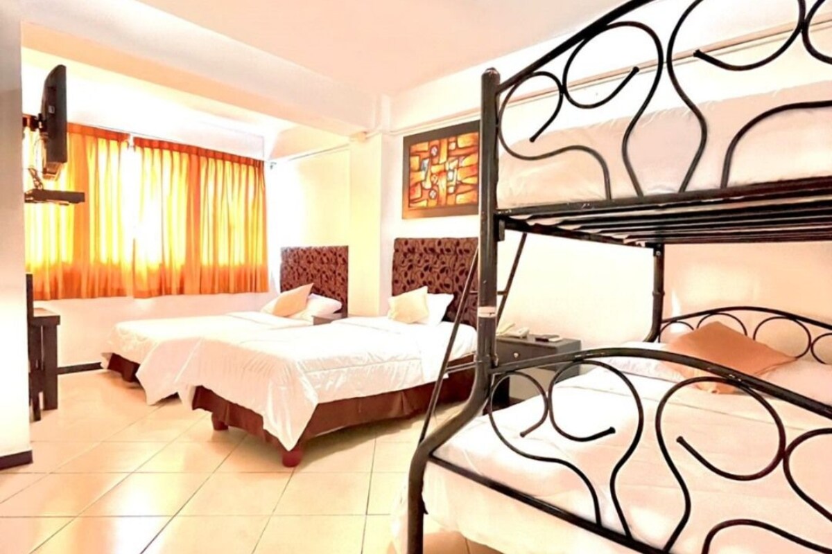 A hotel room with a bunk bed, two single beds, a flat-screen TV, and a large window with curtains.