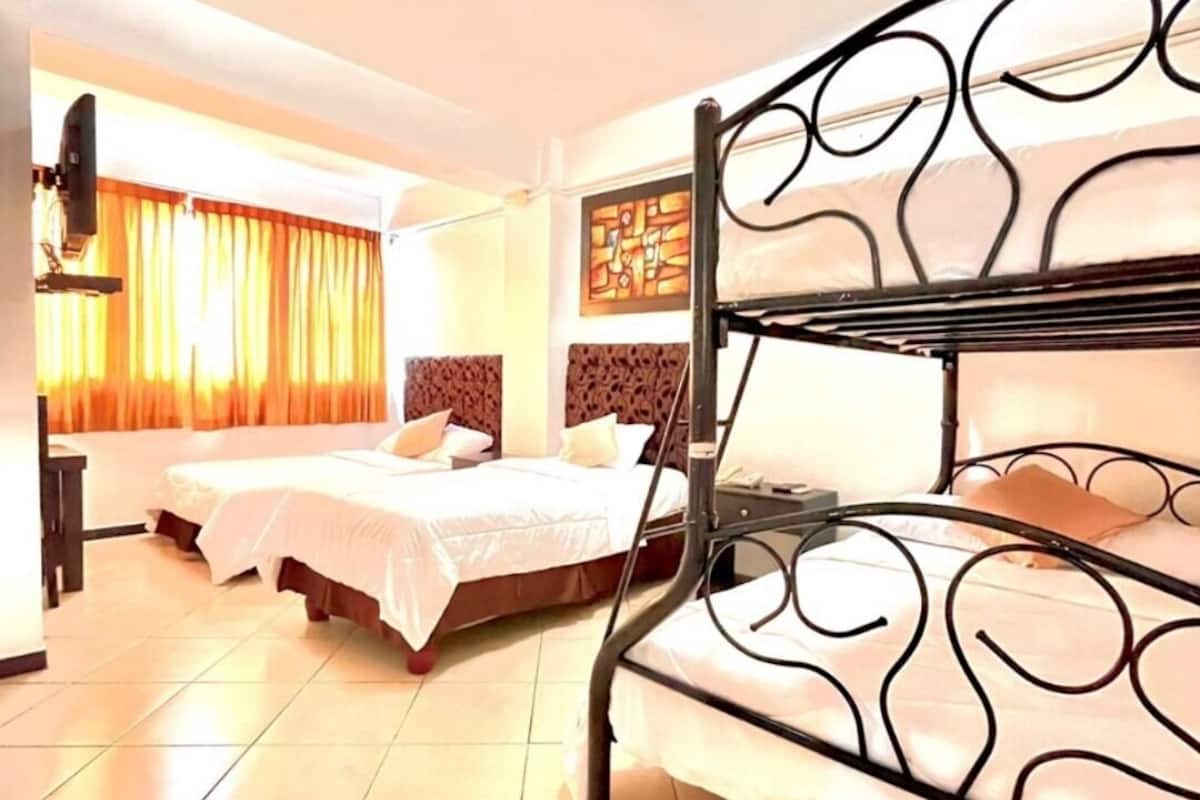 Quarto de hotel com cama de solteiro, duas camas simples, televisão de tela plana e uma janela grande com cortinas.