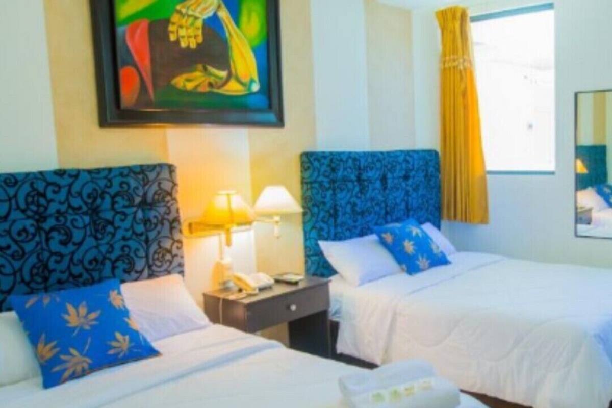 Quarto de hotel com duas camas, cada uma com cabeceira azul e roupa de cama branca. Há um quadro na parede acima das camas, uma janela com cortinas amarelas e um espelho na parede.