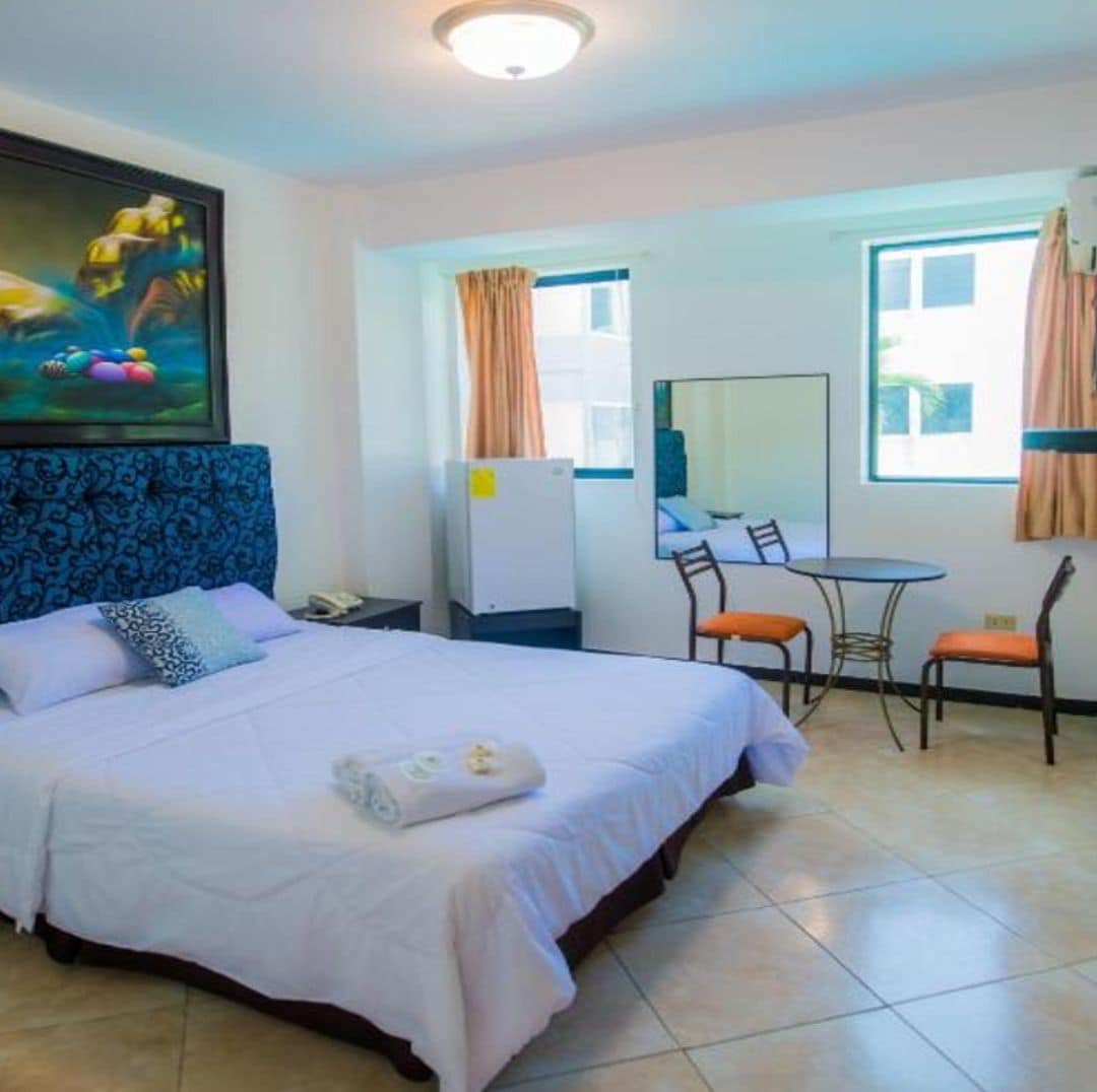A hotel room with a bed, a desk, two chairs, a mirror, and a painting on the wall.