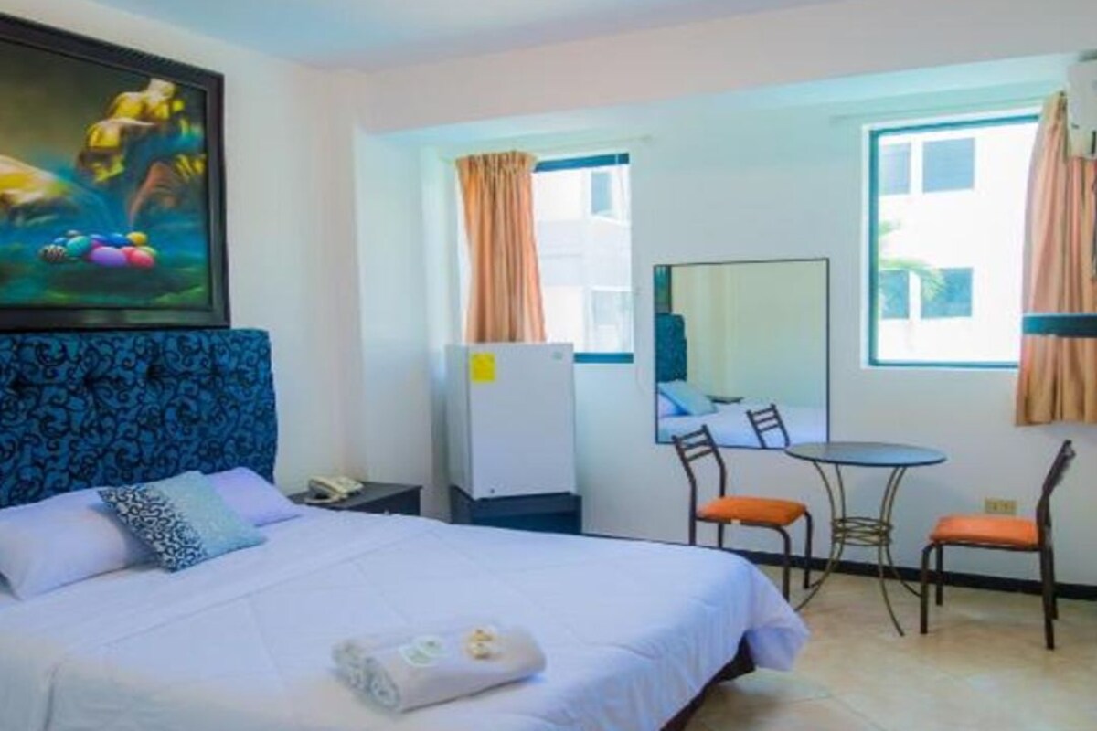 A hotel room with a bed, a desk, two chairs, a mirror, and a painting on the wall.