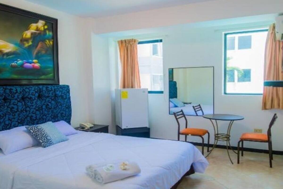 Quarto de hotel com cama, escrivaninha, duas cadeiras, espelho e um quadro na parede.